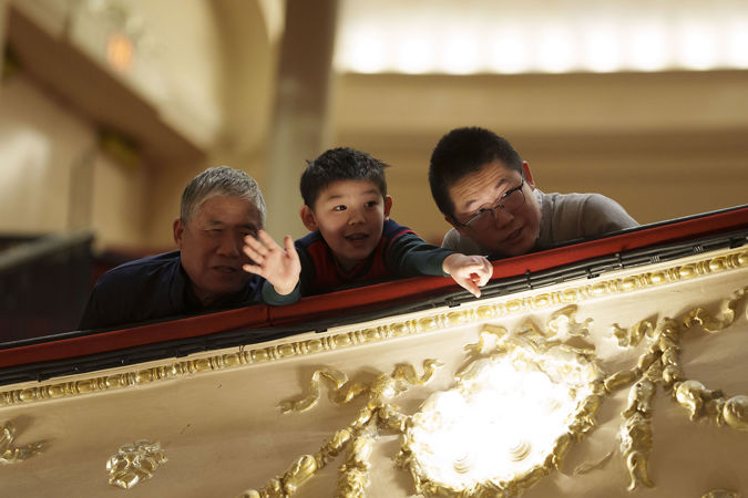 A young audience member pointing towards the stage and two adults looking from the Lower Balcony at the finalists on stage