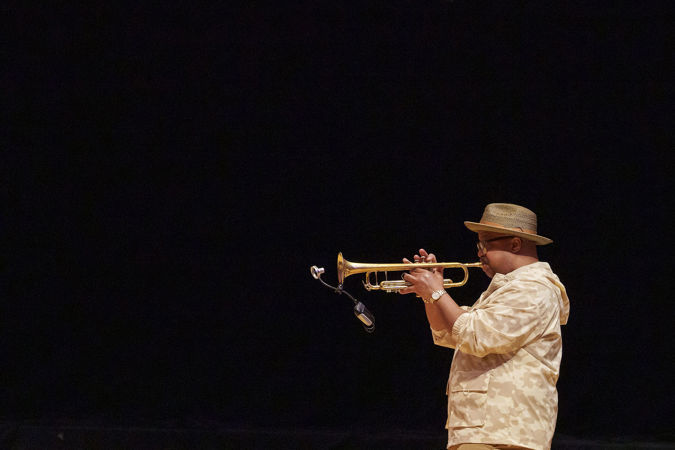 A side profile of Nicholas Payton performing on trumpet. He wears a tan jacket, a brimmed hat and glasses.