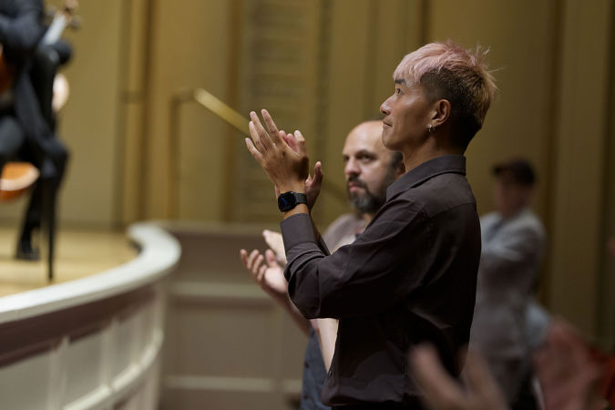 An audience member in the front row, with short pink hair and an elegant black watch, gives the orchestra a standing ovation