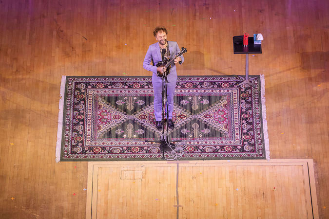 Chris Thile sings and plays mandolin, standing in the middle of an ornate, patterned carpet rolled out for him on stage
